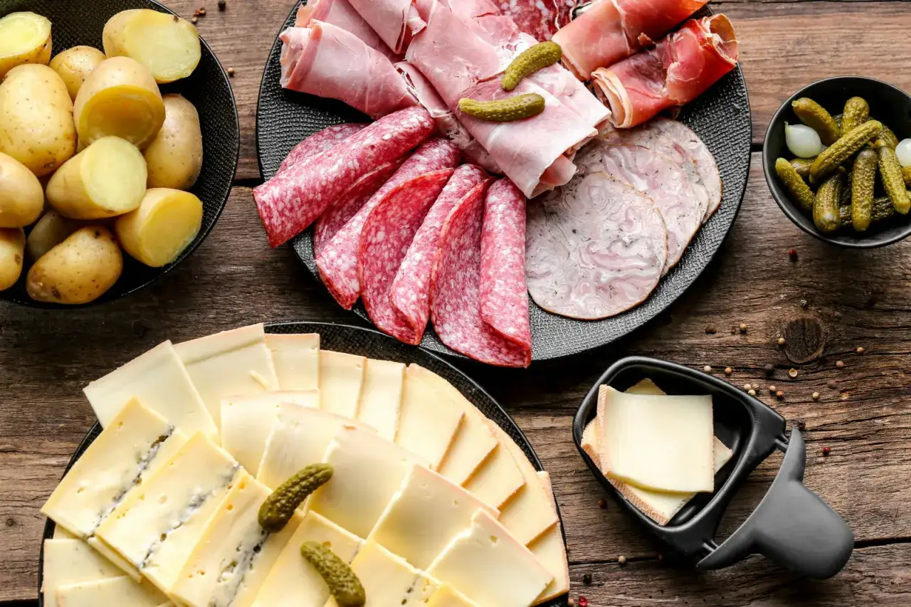 Comment présenter une raclette : plateau de charcuteries, pommes de terre, fromages variés et cornichons sur fond de bois.